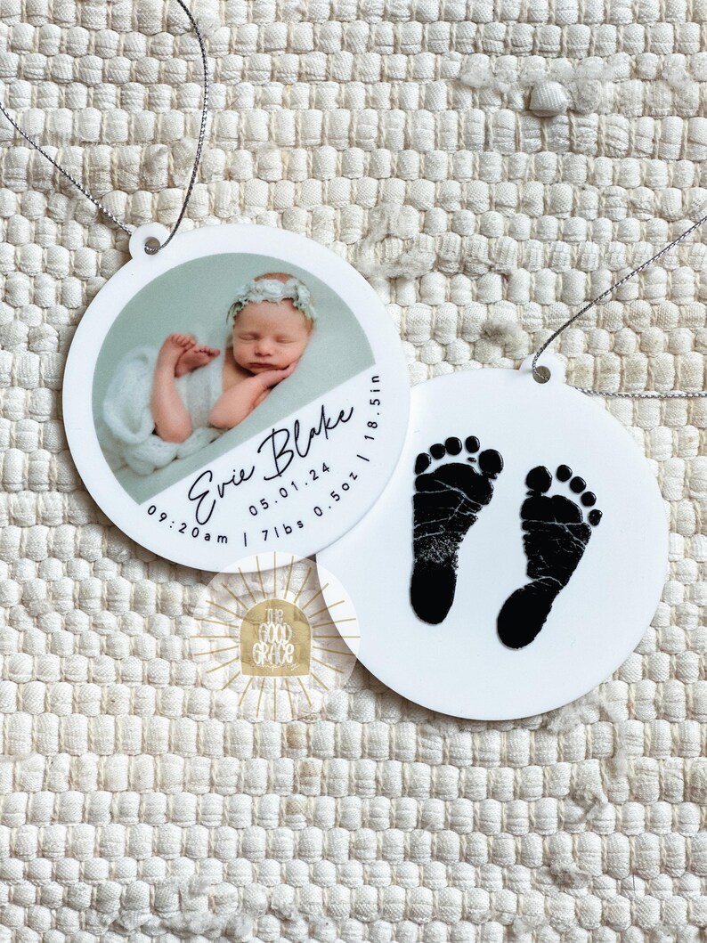 May include: Two round white ornaments with silver hanging strings. One ornament features a photograph of a newborn baby wearing a floral headband, with the text Evie Blake, 05.01.24, 09:20 am, 7 lbs 0.5 oz, and 18.5 in. The other ornament displays black baby footprints.
