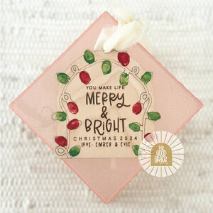 Op de afbeelding: Houten kerstornament met de tekst "You Make Life Merry & Bright Christmas 2024 Love Ember & Evie". Het ornament heeft rode en groene lampjes op een roze, glitterende achtergrond.
