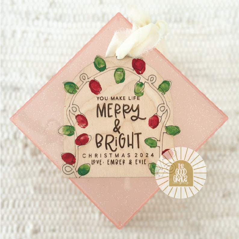 Op de afbeelding: Houten kerstornament met de tekst "You Make Life Merry & Bright Christmas 2024 Love Ember & Evie". Het ornament heeft rode en groene lampjes op een roze, glitterende achtergrond.