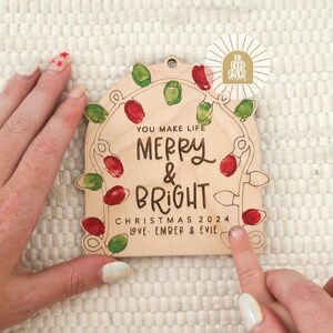 Op de afbeelding: Houten kerstornament met de tekst "You Make Life Merry & Bright Christmas 2024 Love, Ember & Evie." Het ornament heeft een slinger van rode en groene kerstverlichting. Een gouden sticker met de tekst "The Good Grace" staat in de rechterbovenhoek.