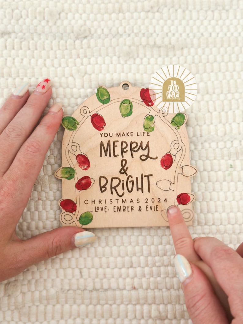 Op de afbeelding: Houten kerstornament met de tekst "You Make Life Merry & Bright Christmas 2024 Love, Ember & Evie." Het ornament heeft een slinger van rode en groene kerstverlichting. Een gouden sticker met de tekst "The Good Grace" staat in de rechterbovenhoek.