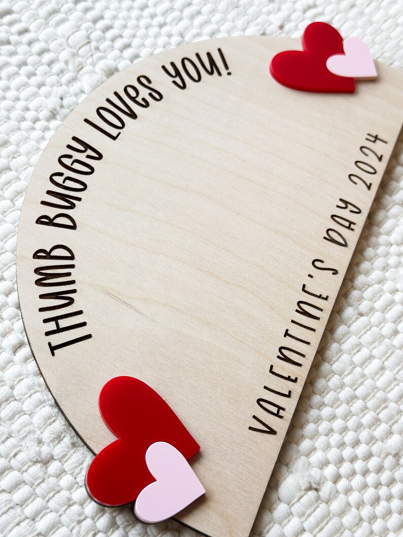 May include: A wooden, semi-circular sign with the text "THUMB BUGGY LOVES YOU!" and "VALENTINE'S DAY 2024" in black. Two sets of layered hearts, one red and one pink, are affixed to the wooden sign. The sign is on a white, textured surface.