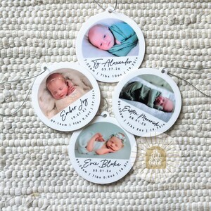 May include: Four circular white ornaments featuring photographs of newborns. Each ornament displays a baby's name, birth date, time, weight in lbs and oz, and length in inches. Names include Ty Alexander, Ember Joy, Easton Maverick, and Evie Blake.