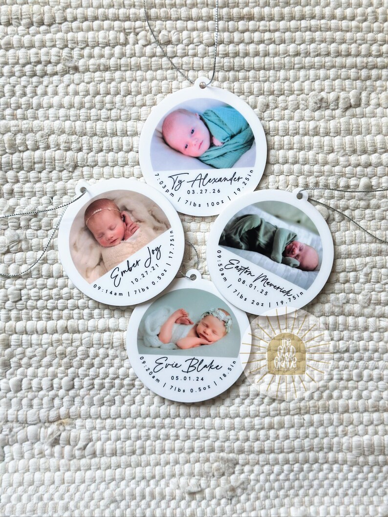May include: Four circular white ornaments featuring photographs of newborns. Each ornament displays a baby's name, birth date, time, weight in lbs and oz, and length in inches. Names include Ty Alexander, Ember Joy, Easton Maverick, and Evie Blake.