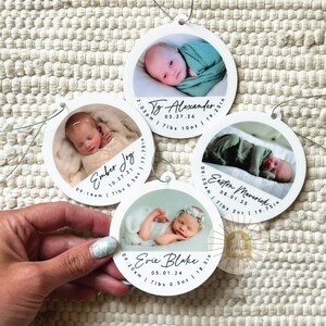May include: Four circular white ornaments featuring photographs of newborns. Each ornament displays a baby's name, birth date, time, weight in lbs and oz, and length in inches. The ornaments have silver hanging strings and are arranged on a textured surface.
