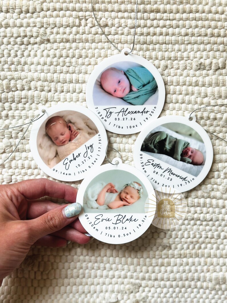 May include: Four circular white ornaments featuring photographs of newborns. Each ornament displays a baby's name, birth date, time, weight in lbs and oz, and length in inches. The ornaments have silver hanging strings and are arranged on a textured surface.