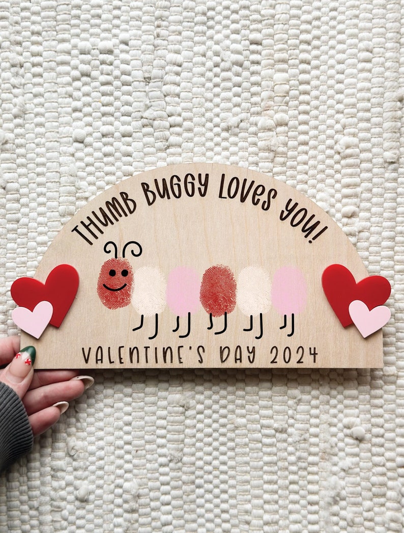 May include: Wooden Valentine's Day sign with the text "THUMB BUGGY LOVES YOU!" and "VALENTINE'S DAY 2024". Features a thumbprint caterpillar design with red and pink hearts. A unique Valentine's Day gift.