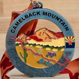 May include: A round medal with the words "Camelback Mountain" in a blue border. The center depicts a desert landscape with a mountain, cactus, and Arizona flag. The medal is attached to an orange ribbon.