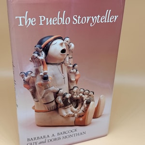 May include: A hardcover book titled The Pueblo Storyteller by Barbara A. Babcock, Guy and Doris Monthan. The cover features a photograph of a ceramic storyteller figurine with a large central figure surrounded by many smaller figures.