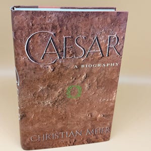 May include: A hardcover book titled Caesar: A Biography by Christian Meier. The front cover features a textured, earthy brown background resembling cracked soil, with the title in large serif font and a small green laurel wreath icon.