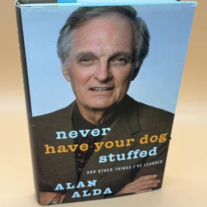 May include: A hardcover book titled "Never Have Your Dog Stuffed" by Alan Alda. The book cover features a portrait of Alan Alda in a brown jacket. The title is in orange and white text.