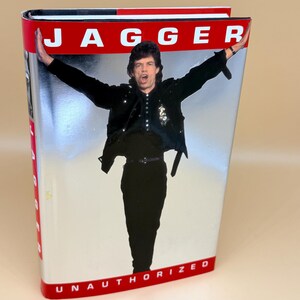 May include: A hardcover book titled Jagger Unauthorized with a silver metallic cover. The front features a photograph of a person with dark hair wearing a black jacket and pants, arms outstretched. The text Jagger appears in white at the top.