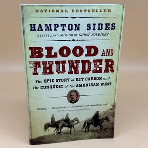 May include: A paperback book titled Blood and Thunder by Hampton Sides. The cover features a sepia-toned photograph of people on horseback, a small portrait of Kit Carson, and the text National Bestseller, The Epic Story of Kit Carson and the Conquest of the American West, and a quote from The Washington Post Book World.