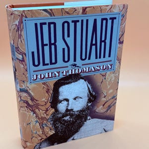 May include: A hardcover book titled "JEB STUART" by John Thomason. The book cover features a portrait of a man with a beard and the title in large, bold letters on a light blue background. The cover has a marbled pattern in shades of brown and cream.