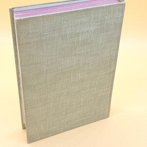 May include: A hardcover book with a light green linen cover. The pages are a soft pink color. The spine is visible with a textured, woven appearance. The book is slightly angled, showcasing its side profile.