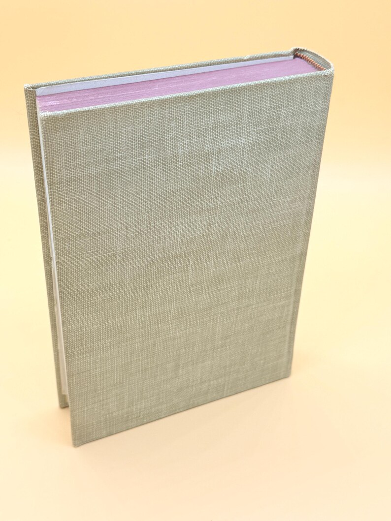 May include: A hardcover book with a light green linen cover. The pages are a soft pink color. The spine is visible with a textured, woven appearance. The book is slightly angled, showcasing its side profile.