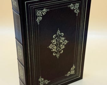 May include: A hardcover book with a dark burgundy cover and gold decorative accents. The cover features an ornate gold design within a rectangular border. A purple ribbon bookmark is visible at the top. The book's spine also has gold detailing.