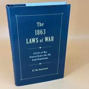 May include: A navy blue book titled "The 1863 Laws of War." The cover features white text within a white rectangular border. The text includes "Articles of War," "General Orders No. 100," and "Army Regulations." The U.S. War Department is also listed.