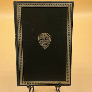 May include: A hardcover book with a dark green cover and gold decorative trim. The book features a gold emblem with the text "VERI TAS" on the front cover. The book is displayed on a black metal stand against a yellow background.