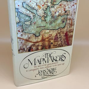 May include: A hardcover book titled The Mapmakers: The story of the great pioneers in cartography-from antiquity to the space age by John Noble Wilford. The cover features a detailed historical map illustration with various flags and landmasses.