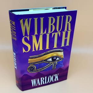 May include: A hardcover book titled "Warlock" by Wilbur Smith. The cover is a deep purple with gold lettering and an illustration of an eye. The book's spine also features the author's name and the title.