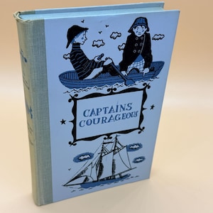 May include: A vintage hardcover book titled "Captains Courageous" with a light blue cover. The cover features illustrations of two boys in a boat and a sailboat. The book's spine is a textured light blue.