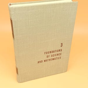 May include: A hardcover book with a light gray linen cover. The title "Foundations of Science and Mathematics" is printed in dark red, along with the number "3". The spine has brown accents.