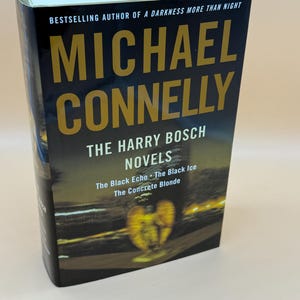 May include: A hardcover book titled The Harry Bosch Novels by Michael Connelly. The black cover features gold text, the author's name, and a graphic of a winged figure against a dark, night-time cityscape. It includes the titles The Black Echo, The Black Ice, and The Concrete Blonde.