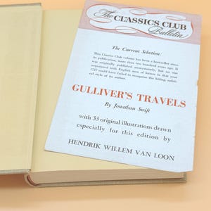 May include: An open book with a beige cover and pages. A white insert with text is placed inside the book. The insert reads "GULLIVER'S TRAVELS" by Jonathan Swift, and "The Classics Club Bulletin".