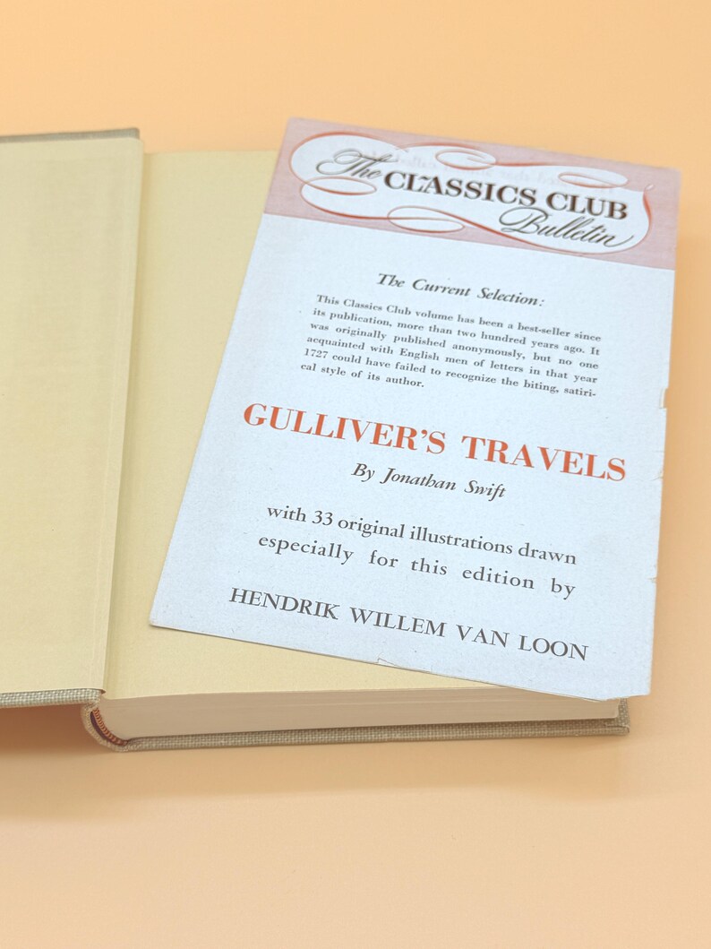 May include: An open book with a beige cover and pages. A white insert with text is placed inside the book. The insert reads "GULLIVER'S TRAVELS" by Jonathan Swift, and "The Classics Club Bulletin".