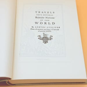 May include: Open book with the title page of "Travels into Several Remote Nations of the World" by Lemuel Gulliver. The text is in black font on a white background, with an ornate emblem below the title. The book is open on a light orange surface.