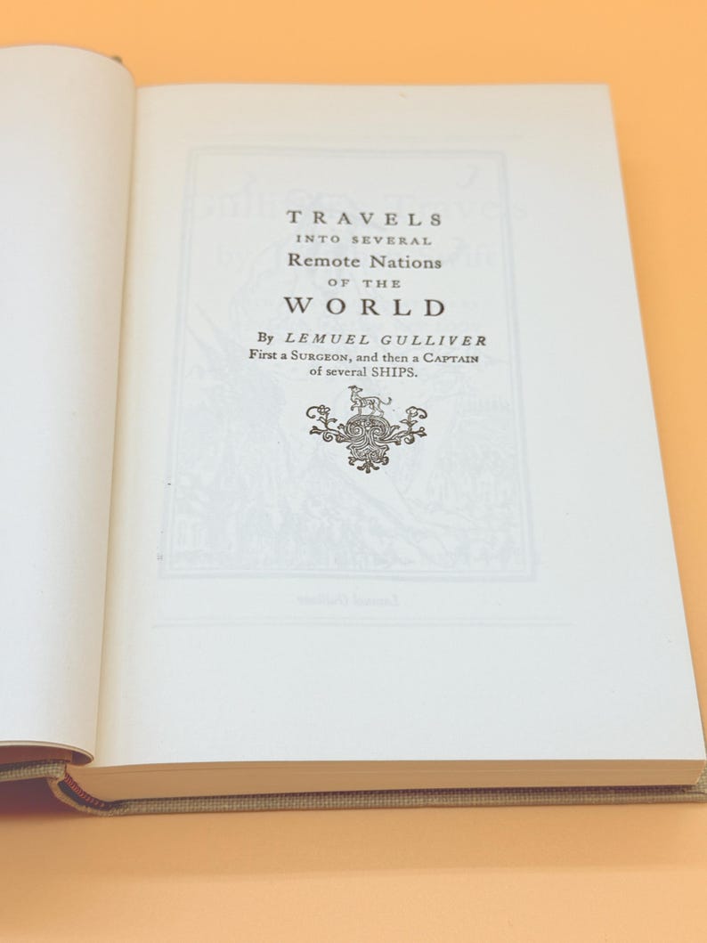 May include: Open book with the title page of "Travels into Several Remote Nations of the World" by Lemuel Gulliver. The text is in black font on a white background, with an ornate emblem below the title. The book is open on a light orange surface.