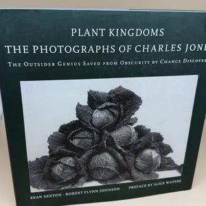 May include: A hardcover book titled Plant Kingdoms: The Photographs of Charles Jones. The dark green cover features white text and a central black and white photograph of a stack of savoy cabbages. Authors are Sean Sexton and Robert Flynn Johnson.