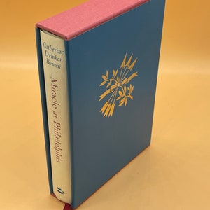 May include: A blue hardcover book with a pink spine. The book title is "Miracle at Philadelphia" by Catherine Drinker Bowen. The cover features a gold illustration of a bouquet of flowers with ribbons.