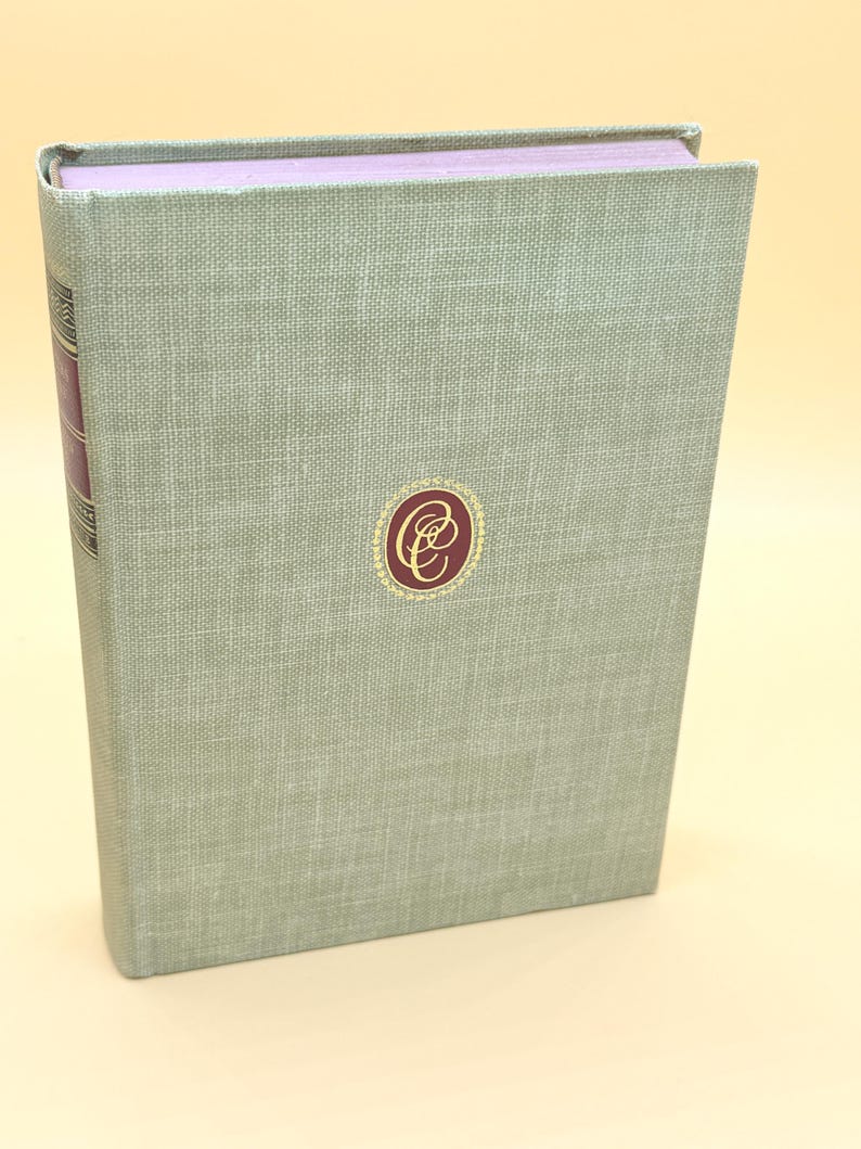 May include: A hardcover book with a light green linen cover. The spine is a darker burgundy color. A gold and burgundy emblem is centered on the front cover. The top edge of the book's pages are a light purple color.