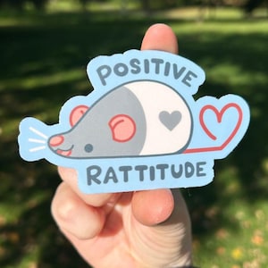 May include: A light blue sticker with the words "POSITIVE RATTITUDE" and a cartoon rat design. The rat has a gray body, pink ears, and a red heart tail. The sticker is held up by a hand against a blurred green background.