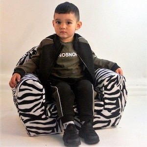 Puede incluir: Un ni&ntilde;o peque&ntilde;o est&aacute; sentado en un sill&oacute;n de estampado de cebra blanco y negro. El ni&ntilde;o lleva una chaqueta negra, una camiseta verde con la palabra "NOSNOH" impresa, pantalones negros y zapatillas negras y blancas.