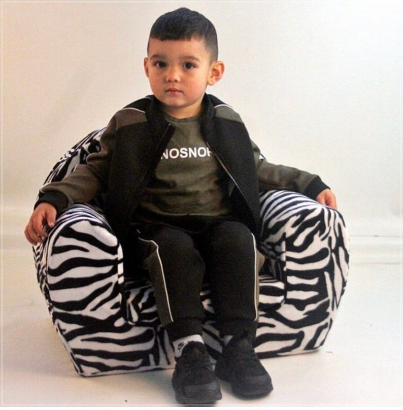 Puede incluir: Un ni&ntilde;o peque&ntilde;o est&aacute; sentado en un sill&oacute;n de estampado de cebra blanco y negro. El ni&ntilde;o lleva una chaqueta negra, una camiseta verde con la palabra "NOSNOH" impresa, pantalones negros y zapatillas negras y blancas.