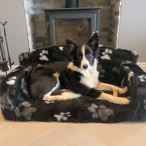 May include: A black and white border collie dog lying on a black and white dog bed with paw print designs. The bed is in front of a fireplace.