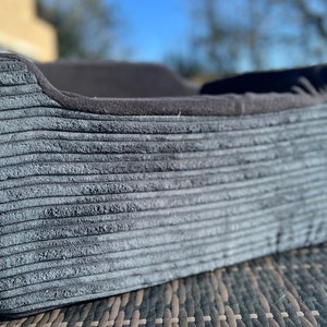 Può includere: Un letto per cani in velluto a coste grigio con un bordo nero. Il letto è appoggiato su una superficie in vimini intrecciato.