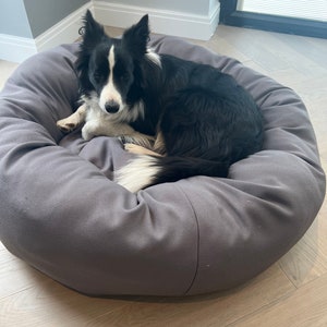 May include: A black and white border collie dog relaxing in a grey round dog bed. The dog bed is on a light wood floor.
