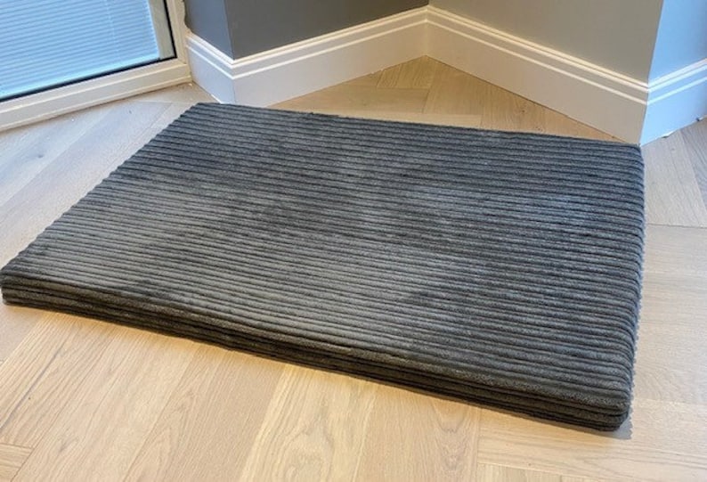 May include: A gray corduroy dog bed with a rectangular shape. The bed is lying on a light wood floor.