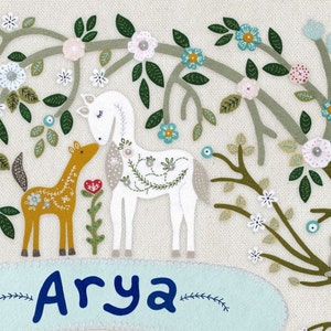 May include: A white horse and a brown horse stand in a field of flowers. The flowers are embroidered in shades of pink, blue, and green. The name "Arya" is embroidered in blue on a light blue background.