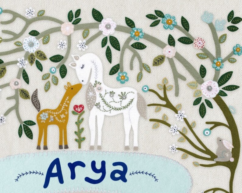 May include: A white horse and a brown horse stand in a field of flowers. The flowers are embroidered in shades of pink, blue, and green. The name "Arya" is embroidered in blue on a light blue background.