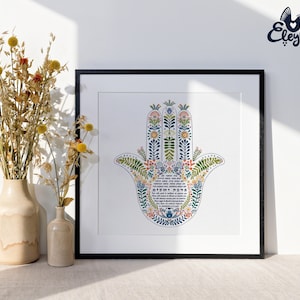 K&ouml;nnte beinhalten: Ein gerahmter Druck mit einem farbenfrohen Hamsa-Hand-Design, verziert mit floralen Mustern in Blau, Gr&uuml;n und Orange. Das Kunstwerk ist vor einem wei&szlig;en Hintergrund und in Schwarz gerahmt. Getrocknete Blumen in beigen Vasen sind im Vordergrund.