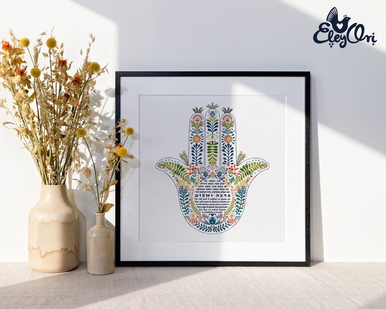 K&ouml;nnte beinhalten: Ein gerahmter Druck mit einem farbenfrohen Hamsa-Hand-Design, verziert mit floralen Mustern in Blau, Gr&uuml;n und Orange. Das Kunstwerk ist vor einem wei&szlig;en Hintergrund und in Schwarz gerahmt. Getrocknete Blumen in beigen Vasen sind im Vordergrund.