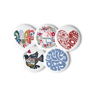 May include: Five round white buttons with colourful designs. The designs include a heart with the words "Bring Them Home", a dove with Hebrew text, a heart with Hebrew text, a floral wreath with Hebrew text, and a circle with Hebrew text.