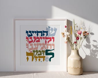 May include: A framed print featuring colorful Hebrew text in shades of blue, red, pink, green, and gold. The artwork is displayed on a white background and framed in a light pink frame. A vase of dried flowers sits to the right of the artwork.