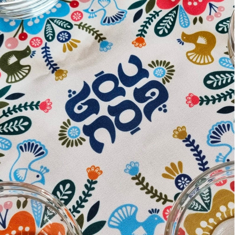 May include: A white fabric with a blue floral pattern and the Hebrew words "Shabbat Shalom" in blue lettering.