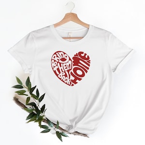 May include: White t-shirt with a red heart-shaped design. The heart is filled with the words "Bring Them Back Home" in white lettering.